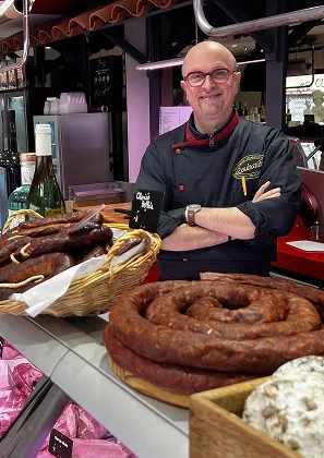 Boucher souriant en uniforme noir, les bras croisés, derrière un comptoir de saucissons à Toulouse en Haute-Garonne 31